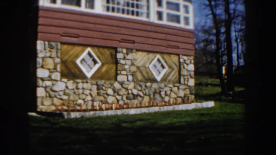 1959 Michigan home-movie frame showing a house with stone foundation, wood siding, diamond windows, grass, and bare trees.