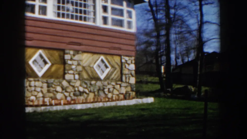 1959 Michigan frame of the same house exterior, with stonework, angled wood panels, diamond windows, and a shaded lawn.