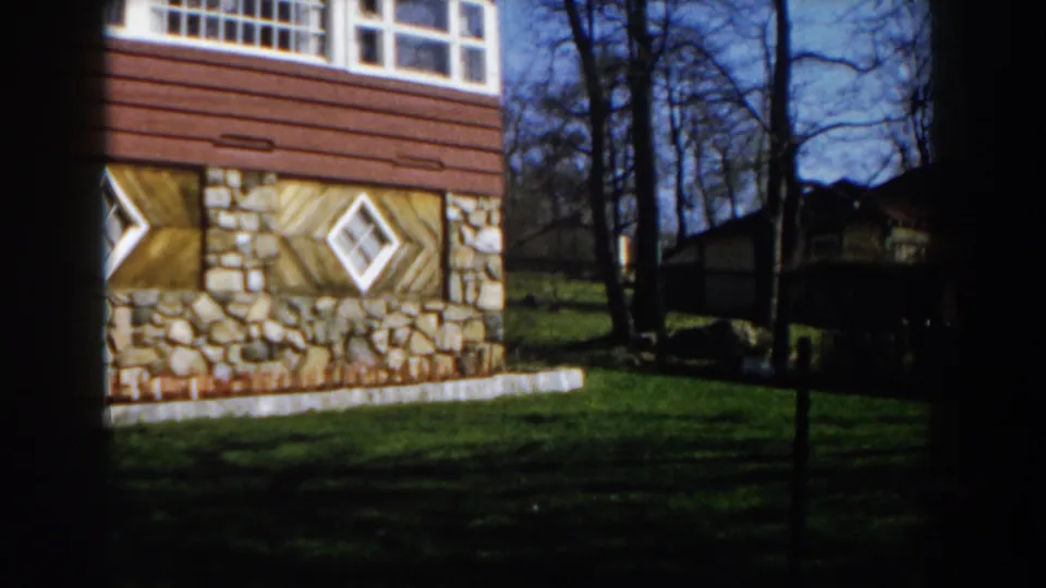 1959 Michigan home-movie view panning past the house corner toward trees and a darker neighboring structure.