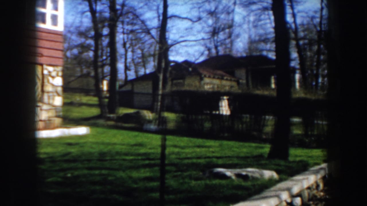 1959 Michigan 8mm frame showing a sunlit lawn, bare trees, stone edging, and residential houses beyond the yard.