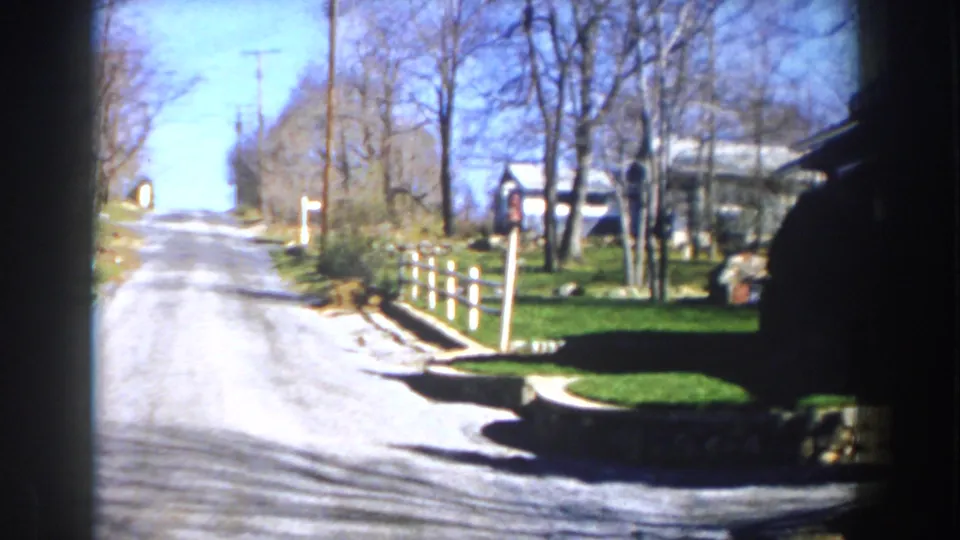 1959 Michigan 8mm frame showing a bright residential road, white fence posts, green grass, bare trees, and houses.