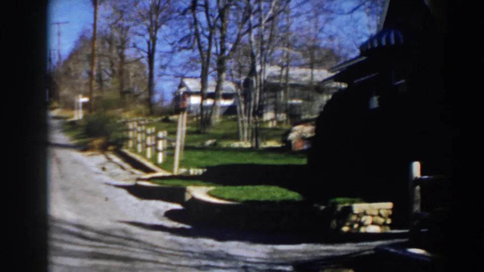 1959 Michigan home-movie frame of a sunny residential street with white fence posts, bare trees, houses, and deep foreground shadow.