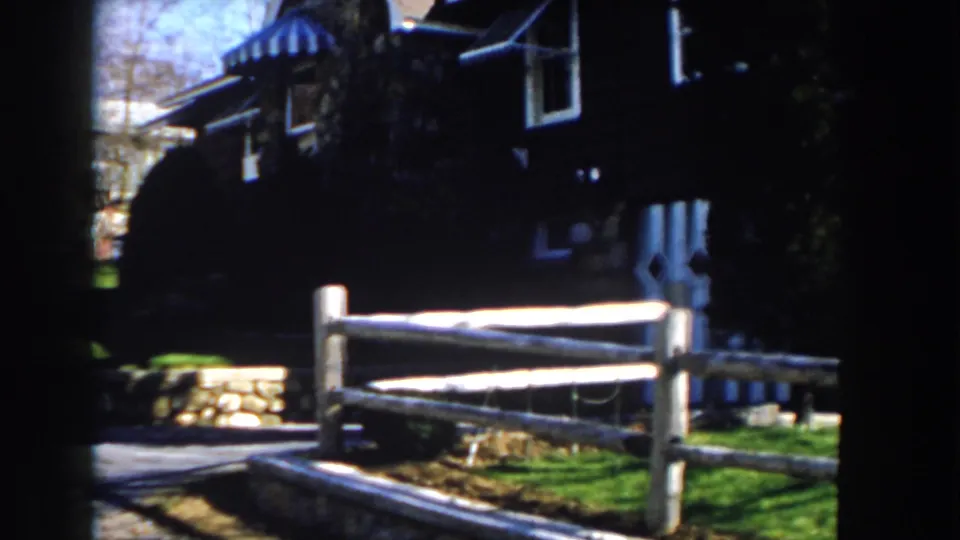 1959 Michigan home-movie frame showing the fence, stone wall, lawn, and shadowed residential exterior during a slow pan.