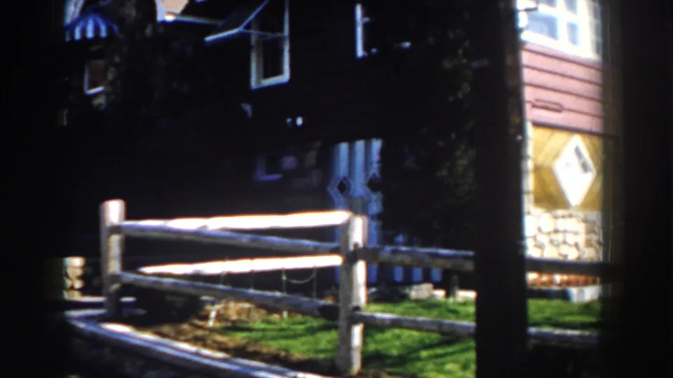 1959 Michigan frame showing a split-rail fence, shaded house exterior, stonework, lawn, and part of a neighboring or adjoining structure.