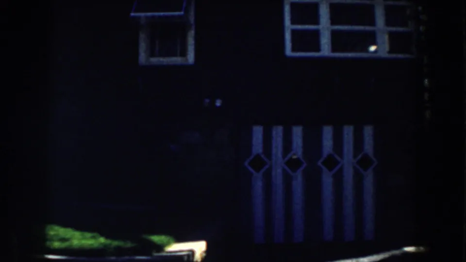 1959 Michigan home-movie frame of a dark cabin wall with multi-pane windows, decorative vertical panels, and a strip of green grass.
