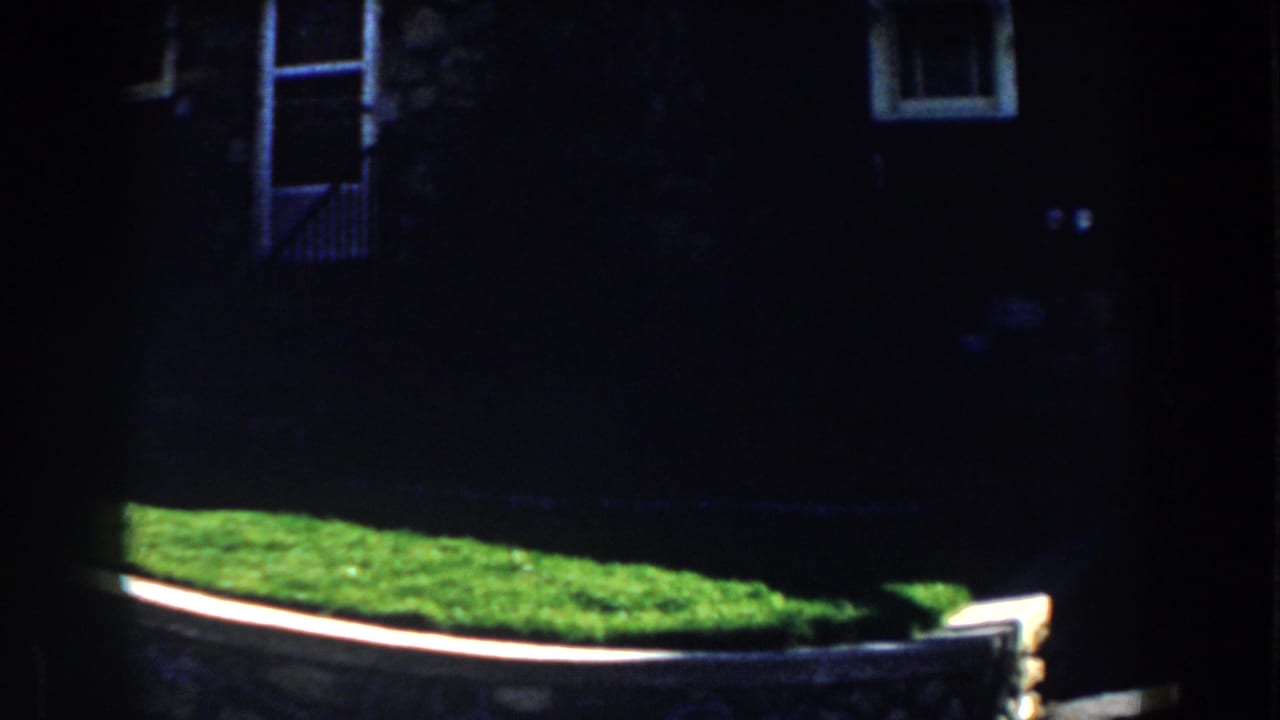 1959 Michigan 8mm frame showing a shadowed stone house exterior, bright lawn, and curved retaining wall.