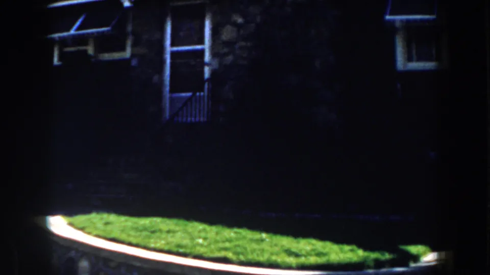 1959 Michigan home-movie frame of a dark stone house facade, upper windows, green lawn, and low retaining wall.