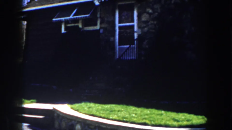 1959 Michigan frame showing a stone house entrance with storm door, small awning, steps, grass, and retaining wall.