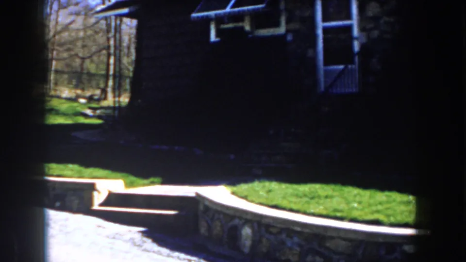 1959 Michigan 8mm frame with a shadowed stone house entry, storm door, porch steps, lawn, retaining wall, and trees beyond.