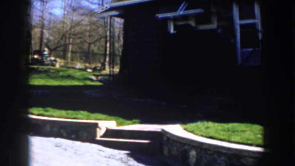 1959 Michigan 8mm frame showing a sunny residential lawn, stone edging, bare trees, and a house mostly in shadow.