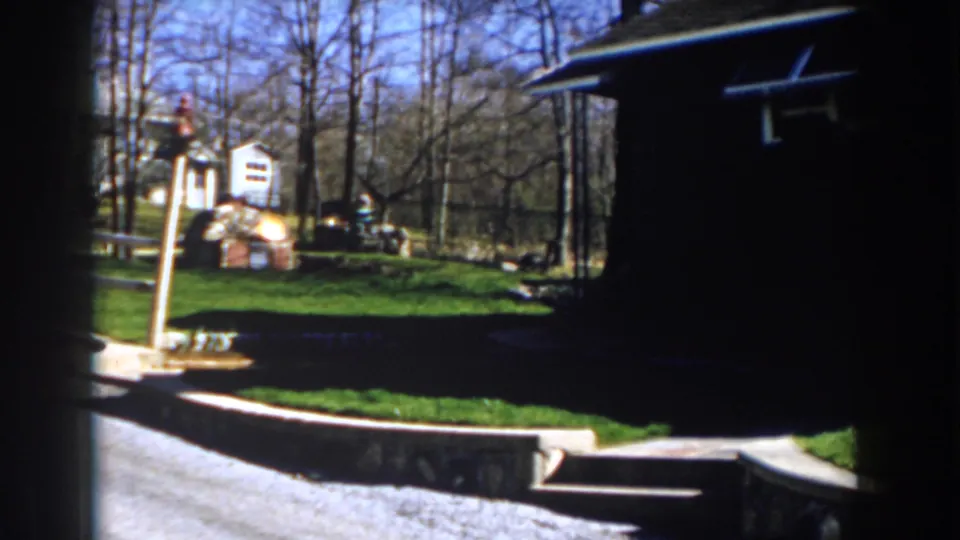 1959 Michigan home-movie frame with a pale wooden fence, small shed, green lawn, bare trees, and outdoor yard items.