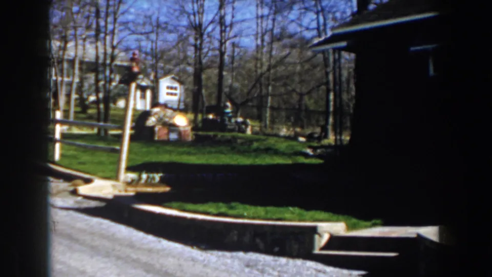 1959 Michigan 8mm frame showing a residential lawn bordered by a wooden fence, with a small shed, stone wall, and bare trees.
