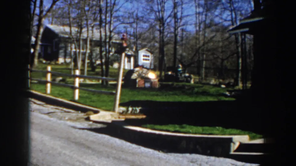 1959 Michigan home-movie frame with a wider view of the lawn, pale fence, small shed, shadowed house edge, and bare trees.