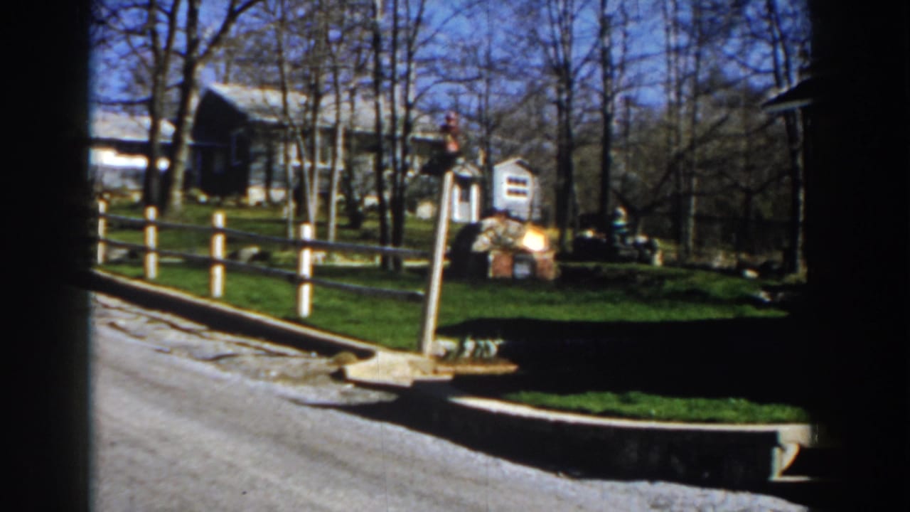 1959 Michigan 8mm frame of a rural road passing a split-rail fence, farmhouse, small mailbox, grassy yard, and leafless trees.