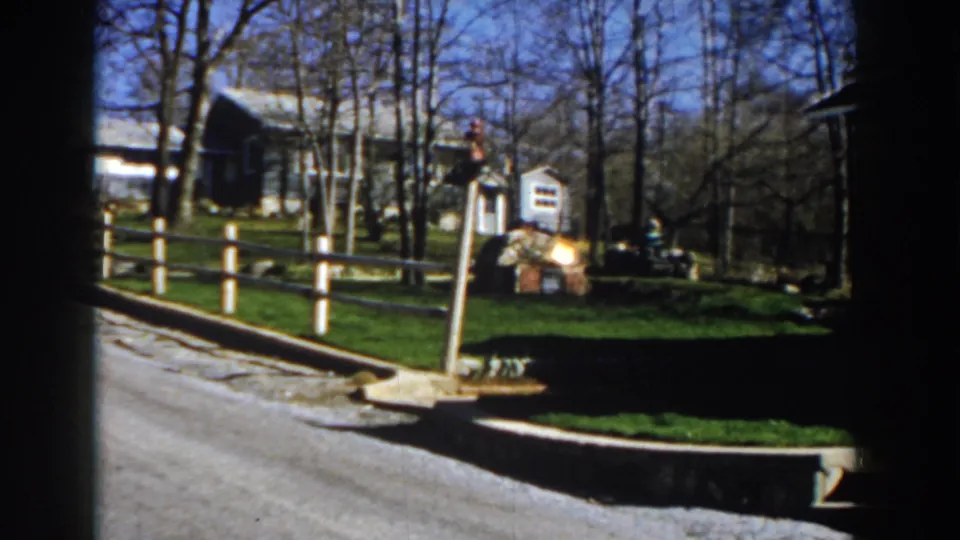1959 Michigan 8mm frame of a rural road passing a split-rail fence, farmhouse, small mailbox, grassy yard, and leafless trees.