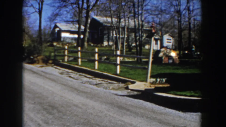 1959 Michigan rural roadside view with a wooden fence, mailbox, farmhouse, lawn, and bare trees seen from a moving vehicle.