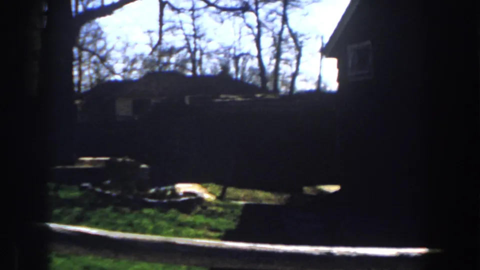 1959 Michigan home-movie frame showing a dim view past a vertical frame toward grass, bare trees, debris, and a building side.
