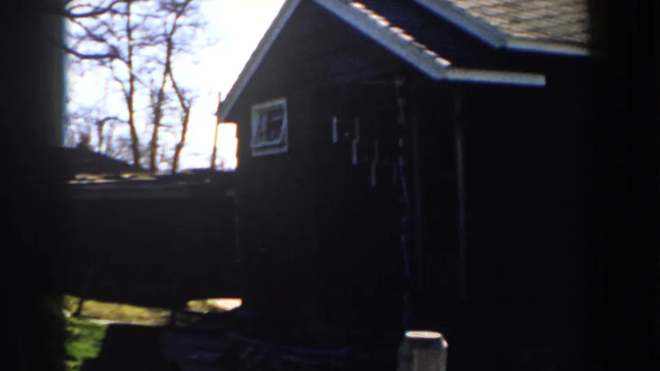 1959 Michigan 8mm frame of a small wood-sided building with a sloped roof, fence line, bare trees, and dark window-edge shadow.