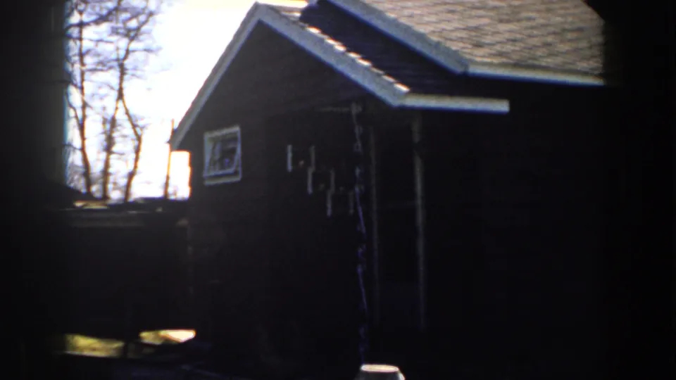 1959 Michigan home-movie view of a small wooden building side, roof eave, window, fence line, and leafless trees beyond.