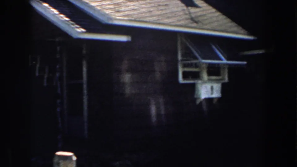 1959 Michigan home-movie frame with a dark house wall, white-trimmed projecting window, and shaded roofline.