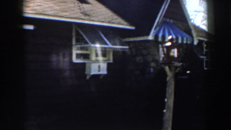 1959 Michigan 8mm frame showing a white-trimmed window, dark siding, striped awning, and yard lamp post.
