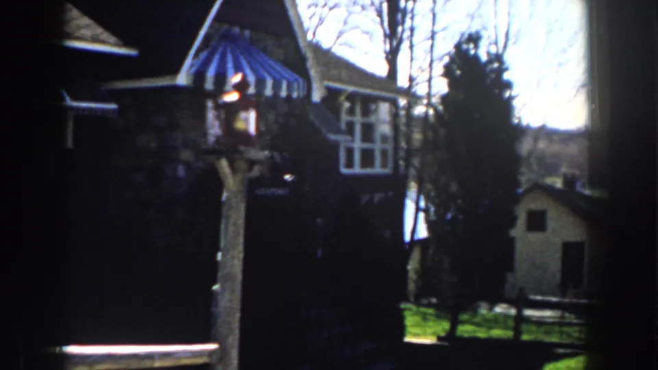 1959 Michigan 8mm frame showing a stone house, blue-and-white striped awning, lantern post, bare trees, and a small shed.