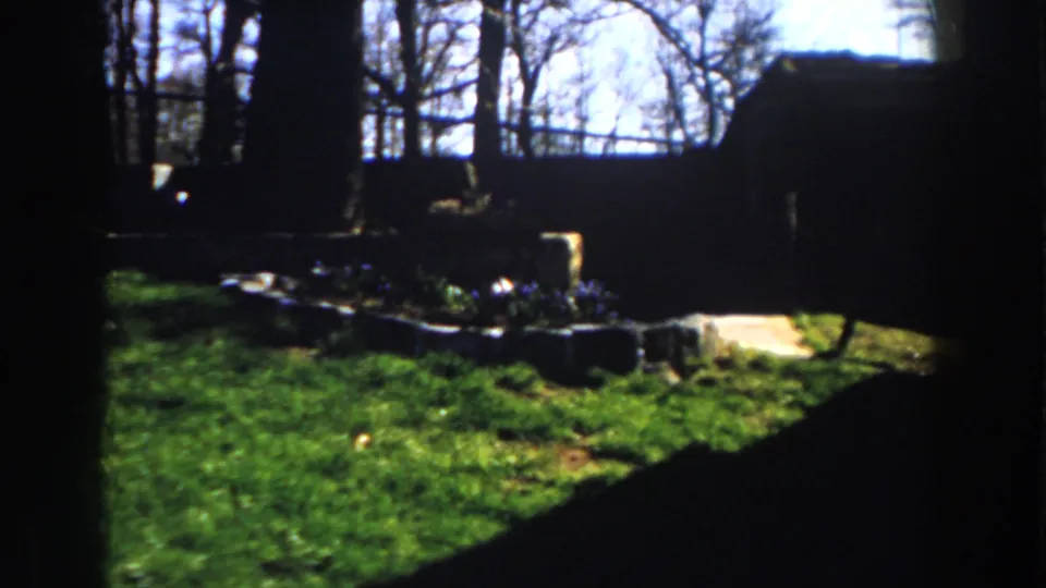 1959 Michigan 8mm frame with a stone-bordered flower bed, lawn, bare trees, and a briefly visible dark figure-like shape at right.