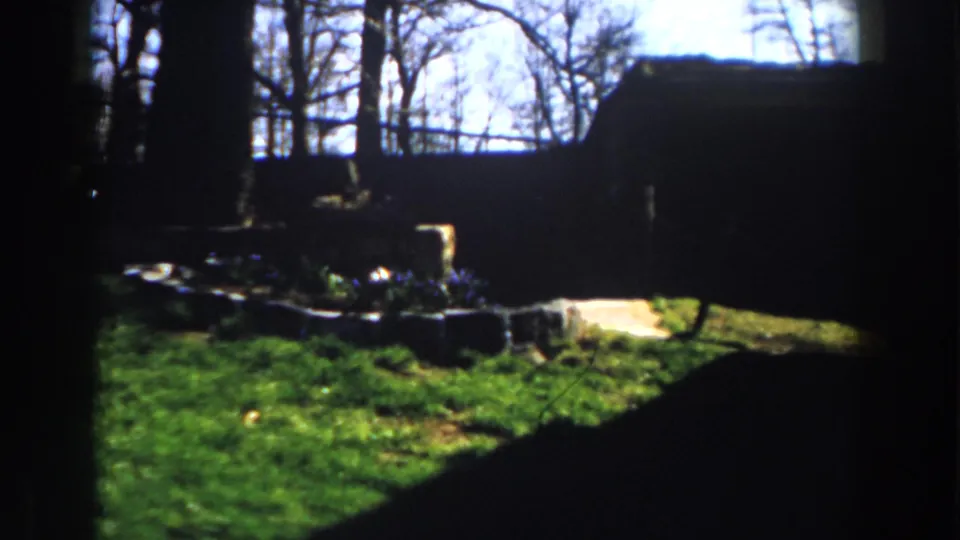 1959 Michigan home-movie frame showing a low stone garden bed, green lawn, bare trees, and deep shadow from a nearby structure.