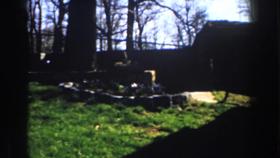 1959 Michigan 8mm frame showing a sunlit backyard from shade, with lawn, bare trees, low stone-edged planting beds, and flowers.