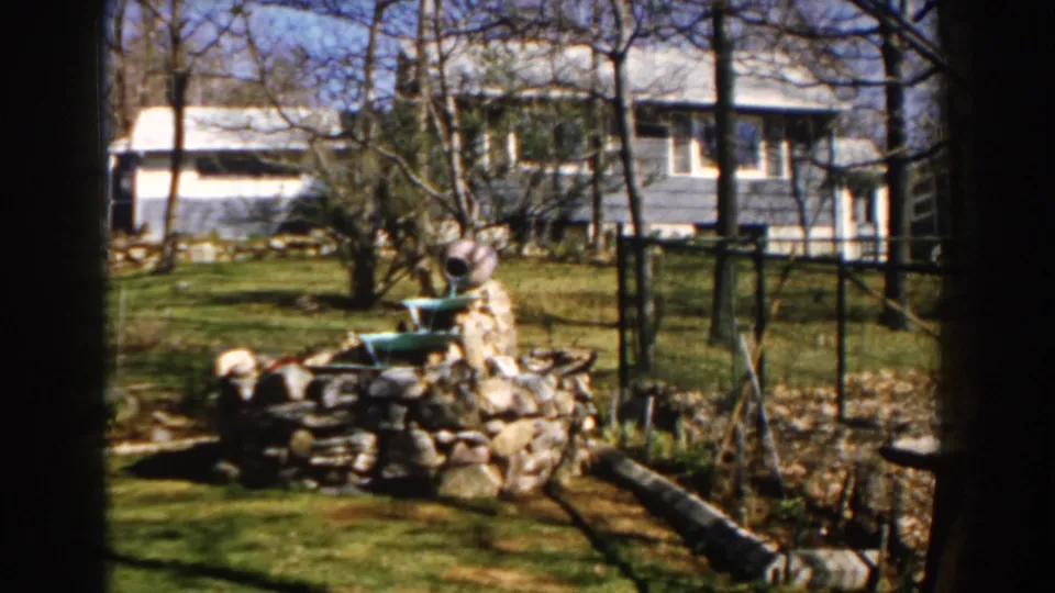 1959 Michigan backyard view with a stone pond feature, ceramic spout, chain-link fence, logs, and blue house beyond.