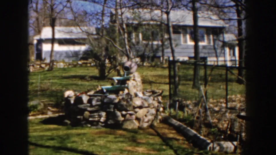 1959 Michigan home-movie frame centered on a stone pond structure with a blue house, fence, logs, and trees in view.