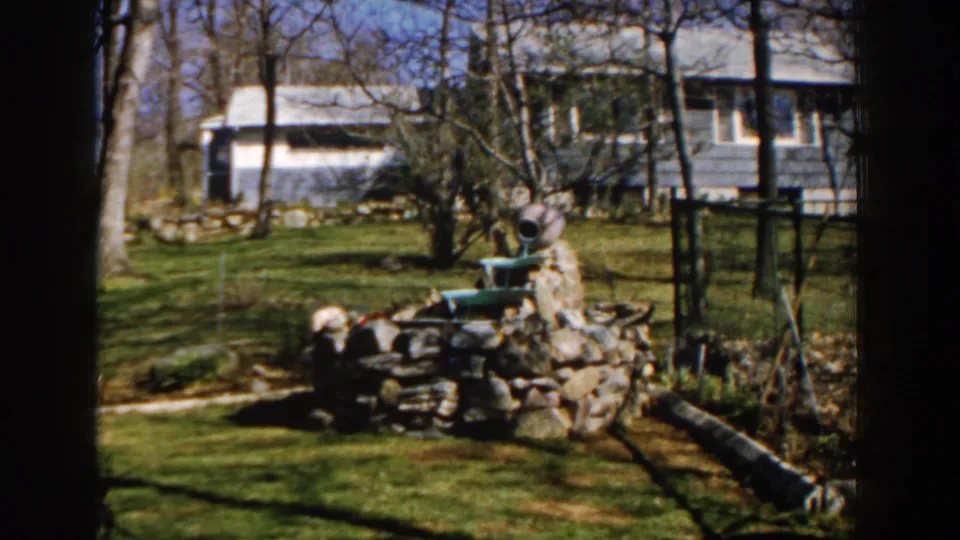 1959 Michigan 8mm frame showing the backyard pond structure, nearby logs, bare trees, chain-link fence, and blue house.