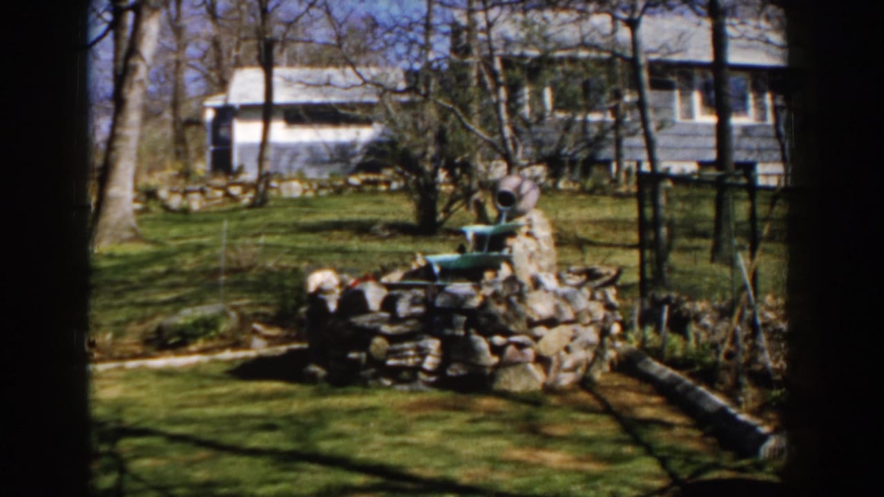 1959 Michigan home-movie frame showing a stone yard fountain with a green basin, bare trees, garden fencing, and a house beyond.