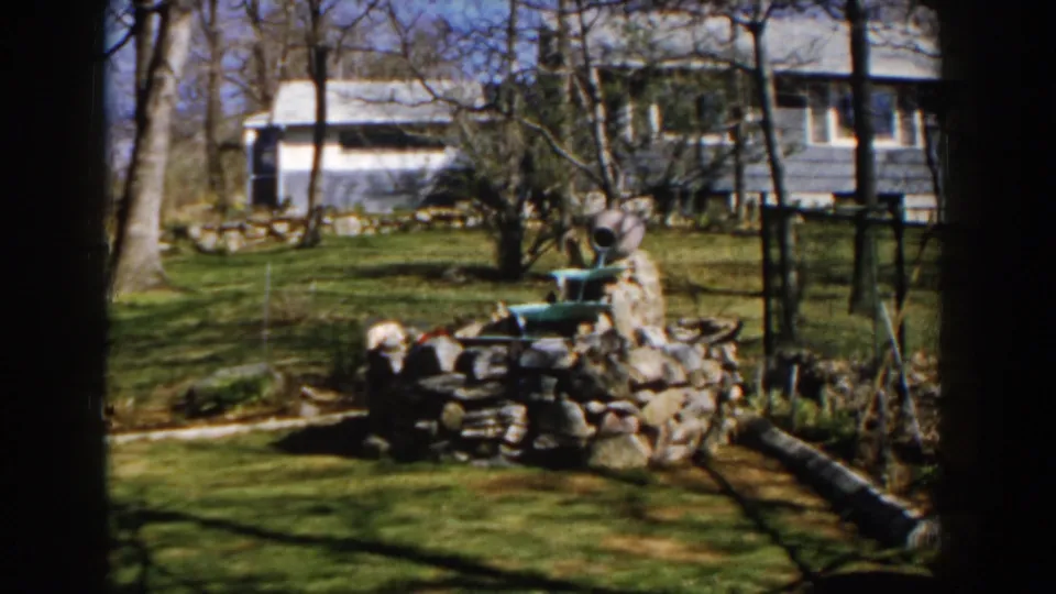 1959 Michigan home-movie frame showing a stone yard fountain with a green basin, bare trees, garden fencing, and a house beyond.