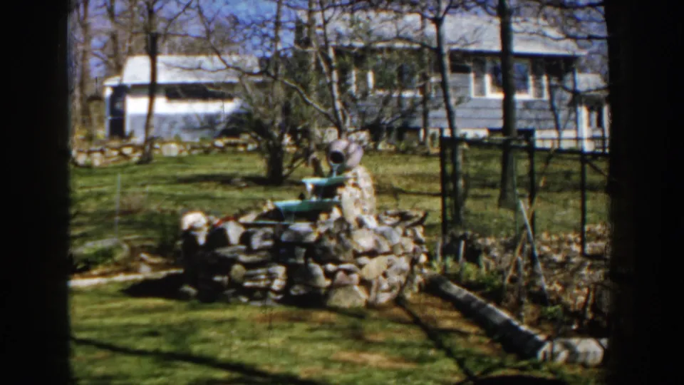 The 1959 Michigan frame shows the stone fountain slightly lower in view, with a green basin, lawn shadows, fence posts, and house siding visible.