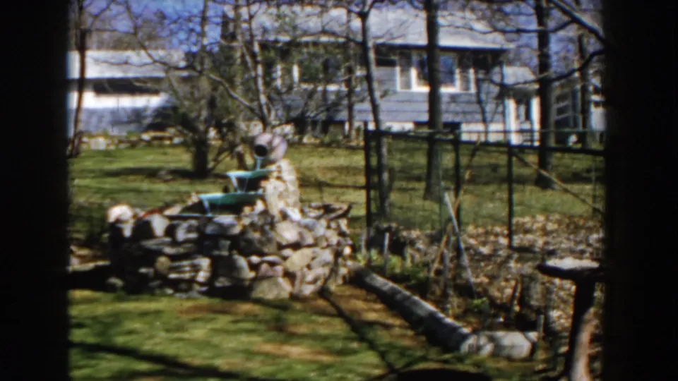 A late 1950s Michigan backyard view holds on the stone fountain, green basin, garden fence, log edging, bare trees, and nearby house.