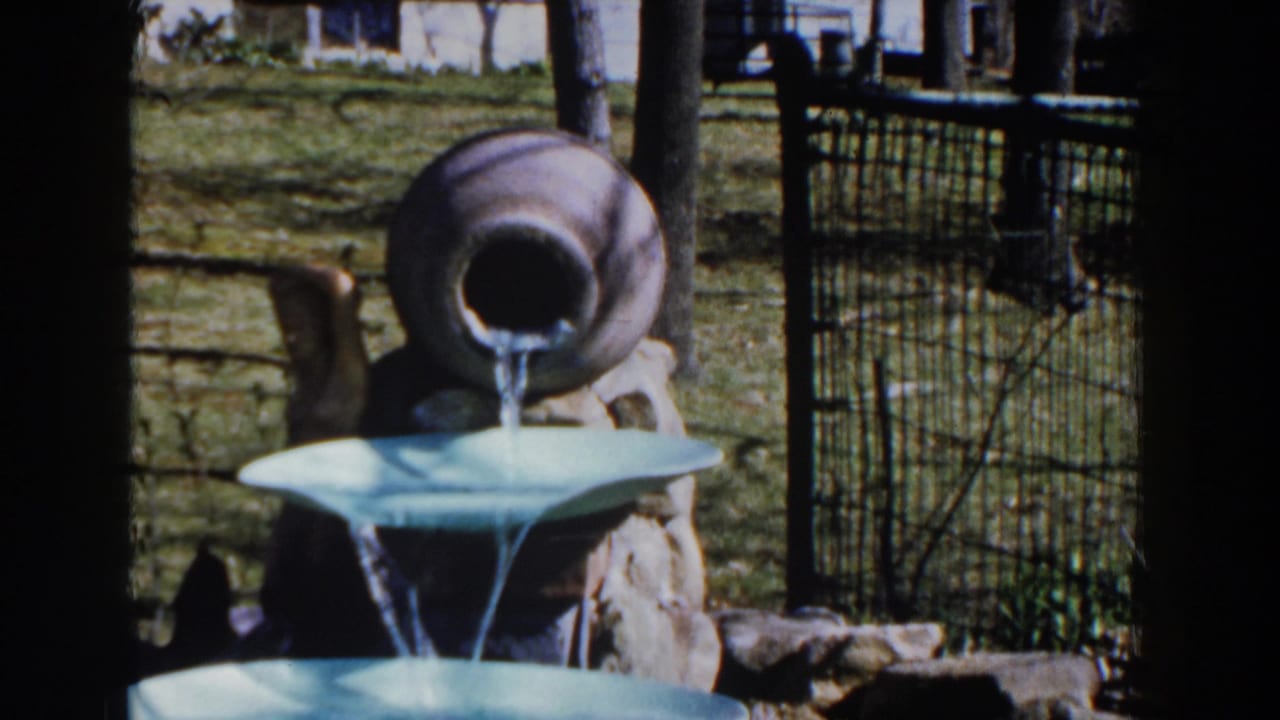 Tan jug pours water into a pale blue tiered fountain in a grassy yard.