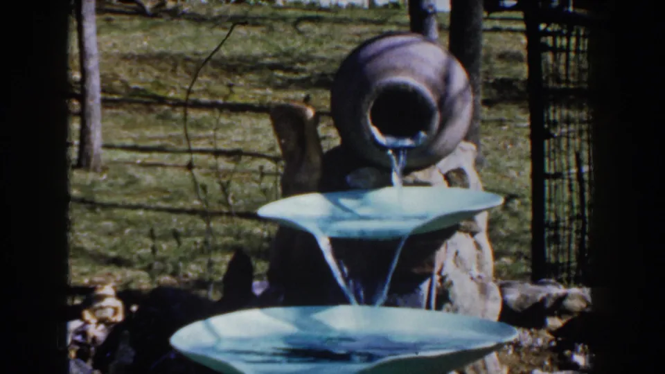 1959 Michigan home-movie frame centered on a tan jug fountain, blue tiered basins, stones, fence, and lawn.