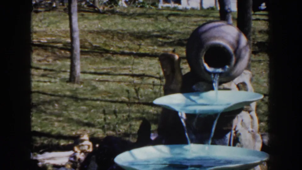 1959 Michigan 8mm frame showing the blue tiered fountain and tan jug against a sunny yard with fence and trees.