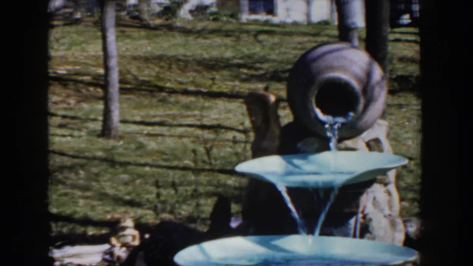 1959 Michigan 8mm frame showing a ceramic jug pouring water into a tiered light-blue garden fountain.