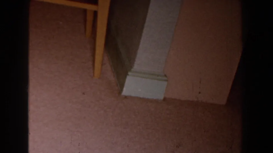 1959 Michigan 8mm frame showing a painted column base beside carpet, a wall corner, and part of a wooden chair leg.