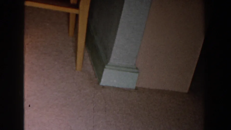 1959 Michigan home-movie frame showing a pale column base on carpet with a wooden chair leg and warm interior wall nearby.