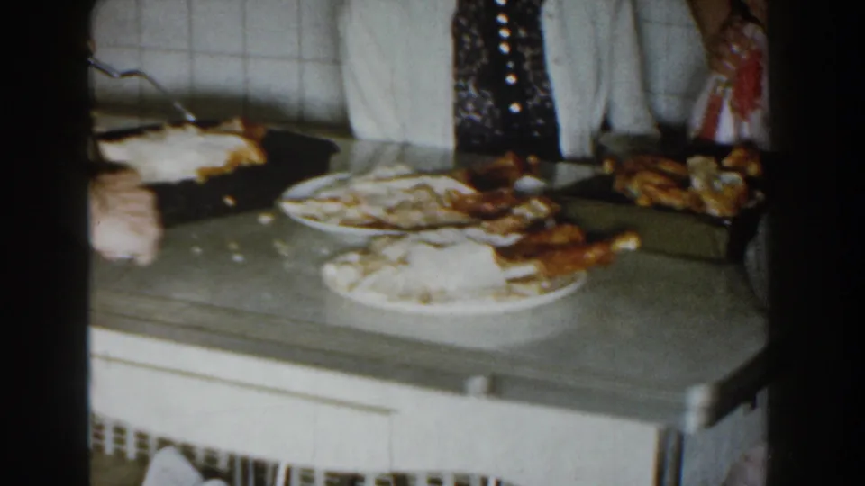 1959 Michigan home-movie frame showing a kitchen counter with plates of food, bread or rolls, and adults partly visible around the serving area.