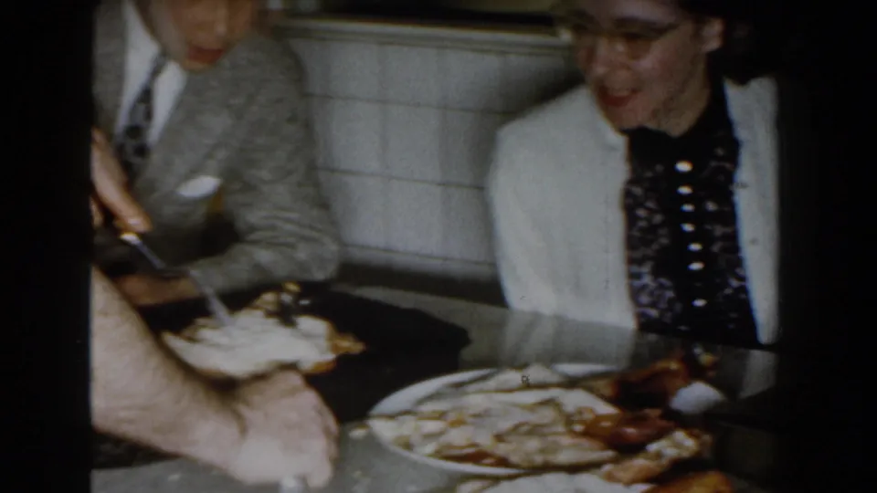 1959 Michigan kitchen frame with hands near serving plates, fried-looking food on the counter, and a woman in glasses smiling at the side.