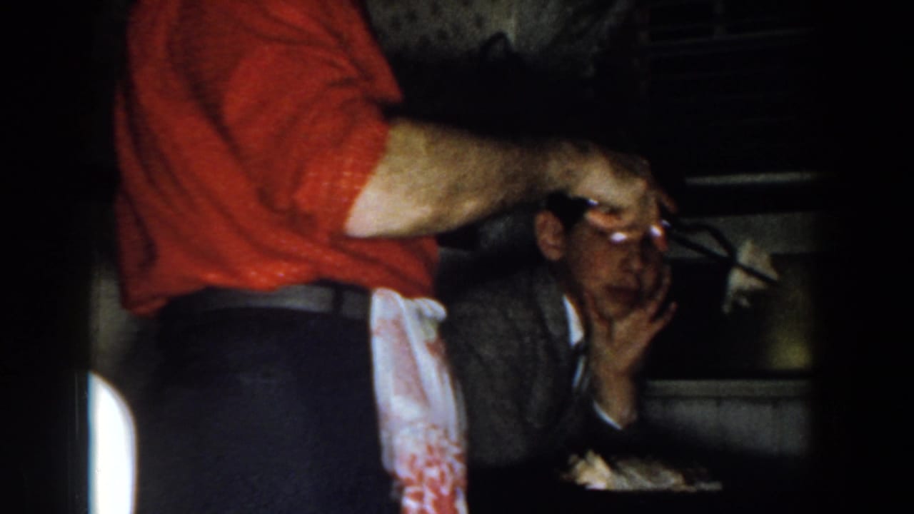 Man in a red shirt and apron serves food near a seated man at a table.