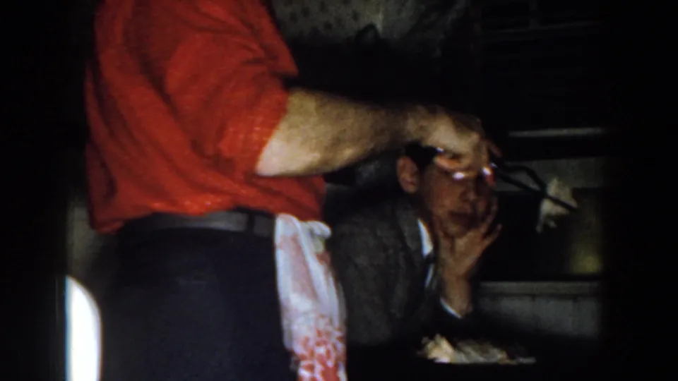 1959 Michigan home-movie frame showing a man in a red shirt and apron serving food beside a seated diner in a dim kitchen interior.
