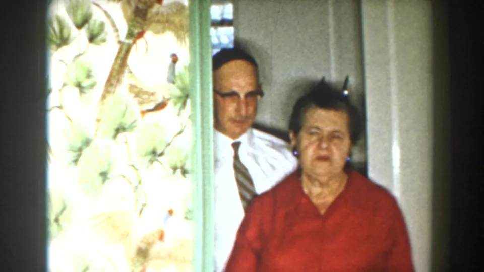 1959 Michigan home-movie frame showing a man in a white shirt and tie beside a woman in red near bird-pattern wallpaper.