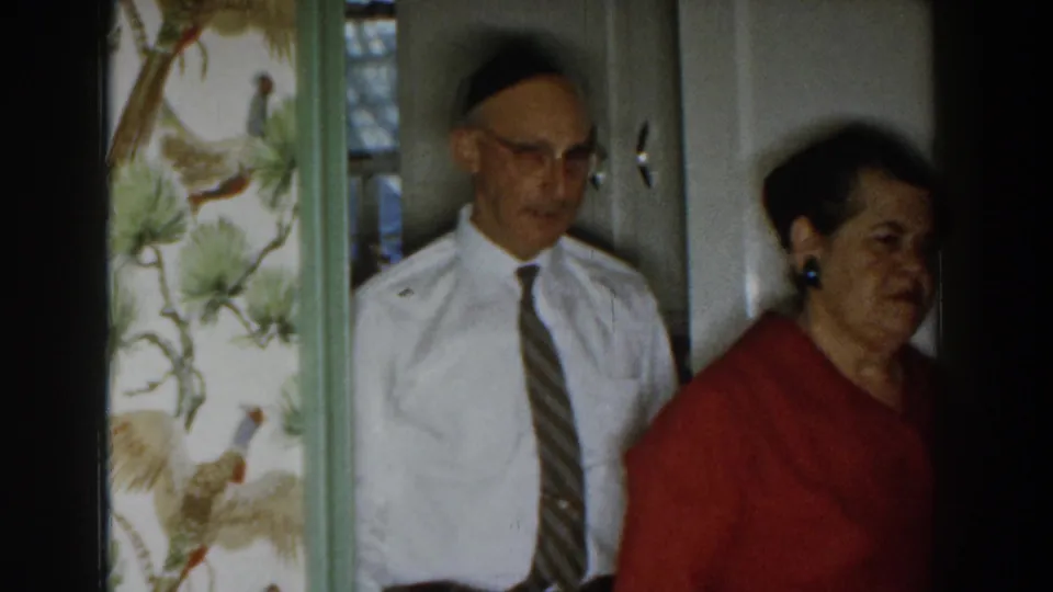 1959 Michigan home-movie frame of two adults standing together inside a home, with bird-pattern wallpaper visible beside them.