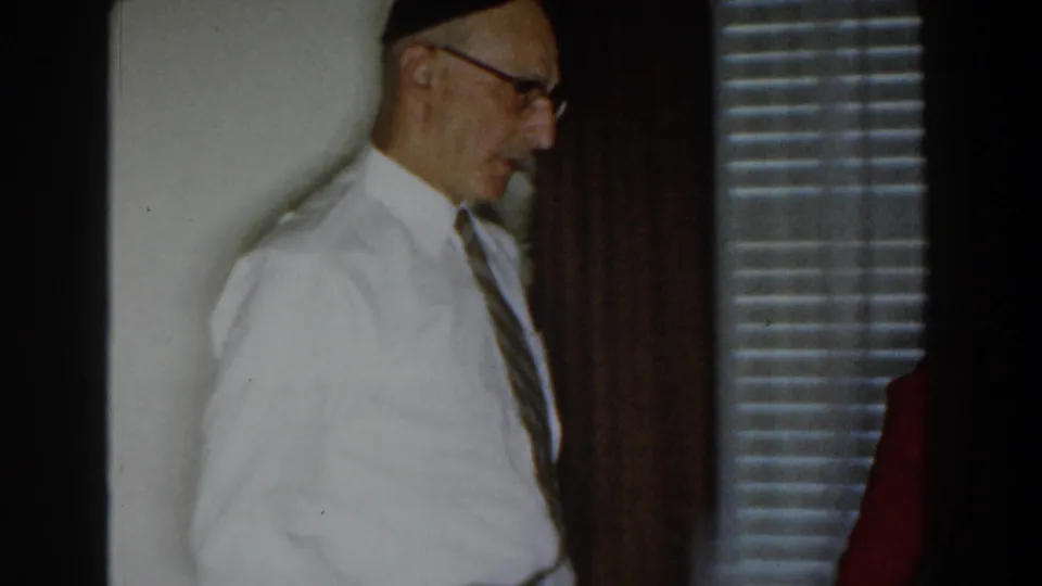 The man in a white shirt and yarmulke moves through a Michigan home interior, with venetian blinds and a red garment at the edge.