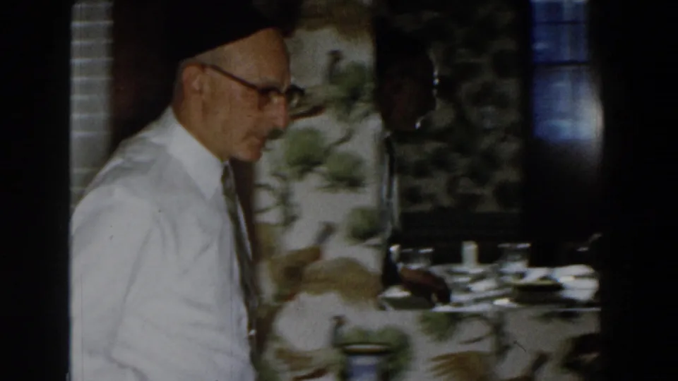 A 1959 Michigan home-movie frame shows the man in profile beside patterned wallpaper, with dishes or tableware visible nearby.